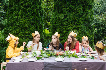 Children wearng paper crowns sitting by  decorated table eating grilled sausages celebrating birthday party in a green garden
