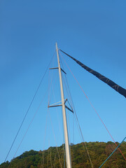 Mast of sailing yacht on a backdrop of clear blue sky and a hill covered with vegetation. Yachting, sailing, water transport, mast without sail. Twisted sail. Close-up, macro, isolated bottom-up view.