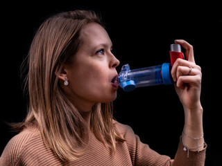 Young women taking asthma medication with an inhaler and spacer with a black background - obrazy, fototapety, plakaty
