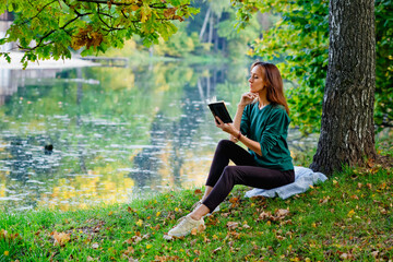 A young, dark-haired woman in a green sweater and trousers sits by a tree at a picnic in autumn park by the lake, holding a book.