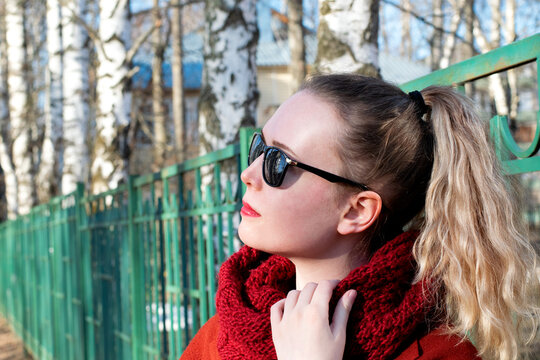 Young blond woman wearing sunglasses, red scarf and with ponytail is standing by the green fence on a sunny day. Spring street style