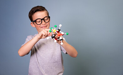 cool schoolboy with black glasses stands in front of blue background holding model of molecules