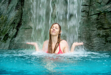 young sexy woman in pink bikini, meditating and enjoy the falling water under the waterfall in the wellness spa pool, copy space