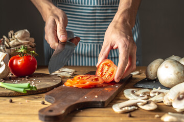 Chef in a blue apron is cutting appetizing fresh tomatoes with a knife to prepare a dish. Concept...