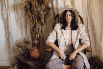 Stylish fashionable woman in suit and hat posing on wooden chair in bohemian room with dry grass