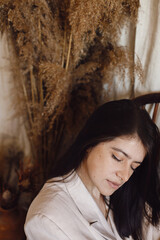Obraz premium Stylish fashionable woman in beige suit posing on wooden chair in bohemian room with dried grass