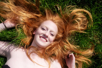 Beautiful portrait of a young sexy red-haired woman, laughing, lying in the spring, summer sun, relaxing on the green meadow grass, the red hair draped freely around the head, Copy space