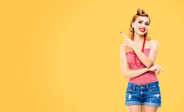 Happy smiling woman pointing at something. Excited girl in pin up, showing product or copy space for text. Retro fashion and vintage. Orange yellow color background.