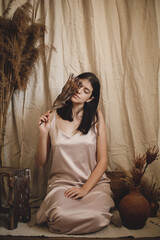 Obraz premium Stylish sensual woman in silk dress holding protea flower at face in bohemian room. Boho bride