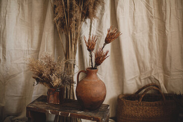 Dry protea flowers in clay vase, pampas grass, wicker basket on background of beige cloth, boho room
