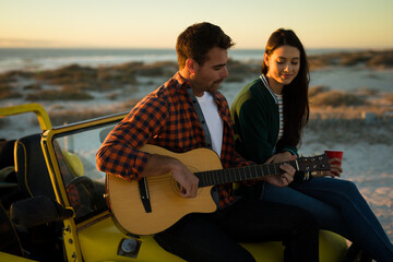 Happy caucasian couple sitting on beach buggy by the sea playing guitar and drinking during sunset