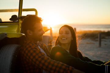 Happy caucasian couple leaning against beach buggy by the sea smiling during sunset