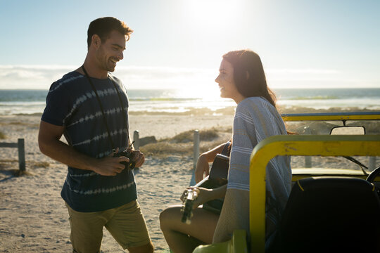 Caucasian couple on beach, man holding camera smiling, woman sitting on beach buggy playing guitar - Powered by Adobe