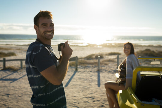 Caucasian couple on beach, man holding camera smiling, woman sitting on beach buggy playing guitar - Powered by Adobe