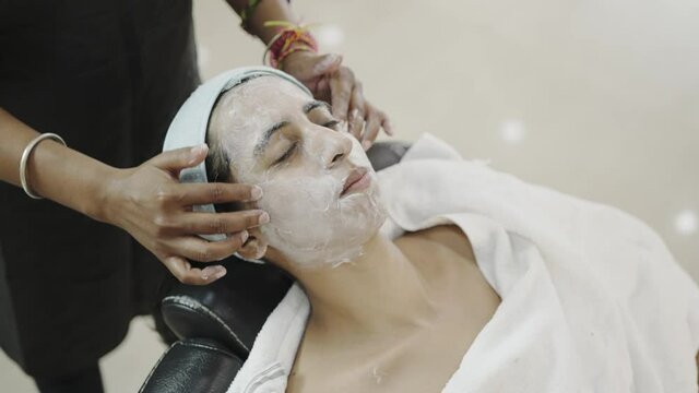close view shot of a lady beautician applying cream along with facial massage on the face of a young Indian Asian female sitting in a comfy chair and receiving a skincare treatment in a beauty parlor