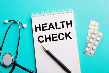 HEALTH CHECK written in a white notepad near a stethoscope, pens and pills on a light blue background. Medical concept