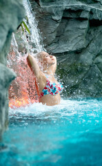 beautiful young cute sexy blonde woman in colorful bikini under the splashing water of the waterfall in the Wellness pool relaxes and enjoys the falling water, copy space