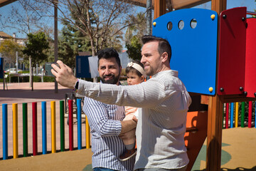 Gay married couple taking a selfie with their cell phone with their daughter in their arms in a playground.