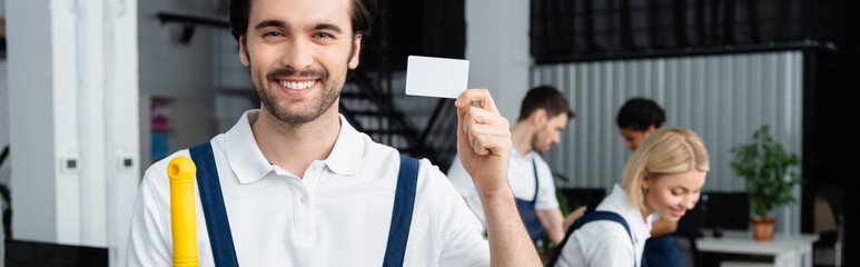 Fototapeta na wymiar Smiling cleaner holding empty card near colleagues in office, banner