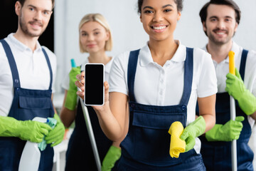 Smartphone in hand of african american cleaner smiling near colleagues on blurred background