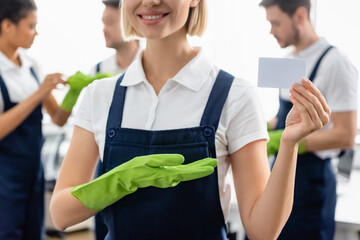 Smiling cleaner pointing at empty card near blurred colleagues in office