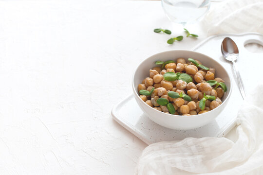 Vegan baked chickpeas with sunflower microgreen sprouts in a bowl on a cutting board on a white textured background