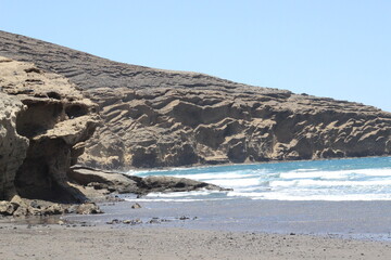 Ocean coast with cliffs. Waves crashing against the brown rocks. Side view of the turquoise ocean  and blue sky. Ocean water and rocks formed from solidified lava of volcano.