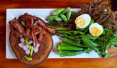 Flat lay of Thai local food with fried or grilled pork, fermented fish, red onions and green chili on wooden cutting board, boiled egg and fresh vegetable on white plate or dish. Spicy Asian food 