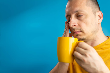 Image of sleepy man 40s with bristle in casual t-shirt drinking coffee while standing isolated over blue background. Morning portrait with cup of coffee. Sleepy guy holding a cup of coffee