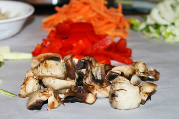 Close-up of chopped mushrooms, red pepper, carrots, and other vegetables laid out for meal preparation. Focus is on mushrooms in the image foreground.