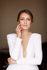 beautiful smiling young bride in long white satin dress with deep neckline, hairstyle and make up standing in hotel room. Trendy wedding style shoot. Wedding day. Bride's morning preparation.
