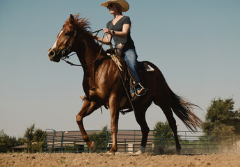 Western lifestyle shows cowgirl riding quarter horse through outdoor arena during summer on ranch.