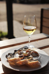 Glass with wine and dish with the snail shells, served on a white classic plate, with various sauces, pesto, truffle, cheese, pieces of French baguette, fork for a shell, wooden table in outdoor cafe