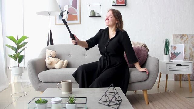young girl in stylish black dress is sitting casual on the gray sofa and talking on the mobile phone on the tripod for phone in hand in light room at home in the morning. lifestyle concept, free space