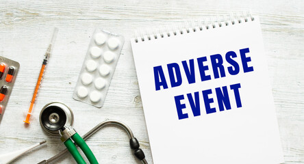 ADVERSE EVENT is written in a notebook on a white table next to pills and a stethoscope.