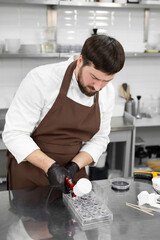 Male pastry chef holds chocolate candy molds and sprays food paint with a pastry airbrush in the kitchen.