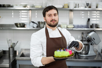 Young smiling chef holds a mousse cake decorated with handmade chocolates.