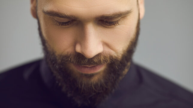 Male face close up. Studio portrait of humble handsome man with moustache and beard in black shirt looking down eyes half closed on plain grey background. People's appearance and human beauty concept