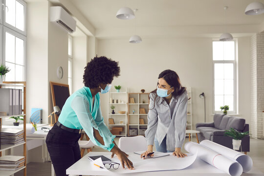Coronavirus pandemic, safety, return to workplace after end of lockdown and quarantine. Two young creative designers and business women in face masks meeting in office studio and working on draft plan - Powered by Adobe