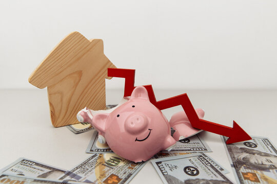 Investment and bankruptcy concept. Real estate. Broken piggy bank and red arrow down with wooden house on dollar banknotes isolated on white