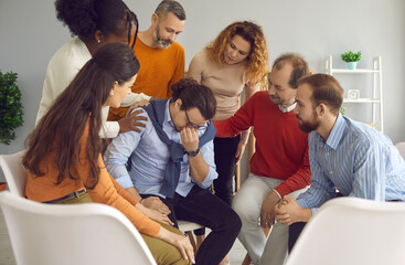 Distressed devastated broken young man suffering from anxiety shares insecurities with people in group therapy. Team of office colleagues comfort and support coworker crying over personal problems