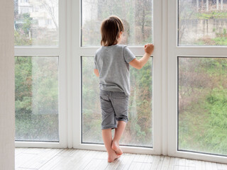Little boy looks outdoors through wet panoramic window with raindrops. Rainy summer weather. Kid stays alone at home. Loneliness and sad mood without children's company.
