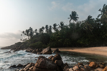 Tropikalna plaża, palmy oraz skaliste wybrzeże na tle zachodzącego słońca.