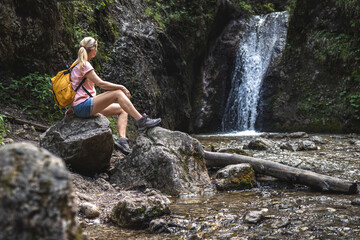 Woman resting during hike in wonderful nature. Hiker sitting on rock near stream with waterfall in forest at natural parkland Mala Fatra, Slovakia