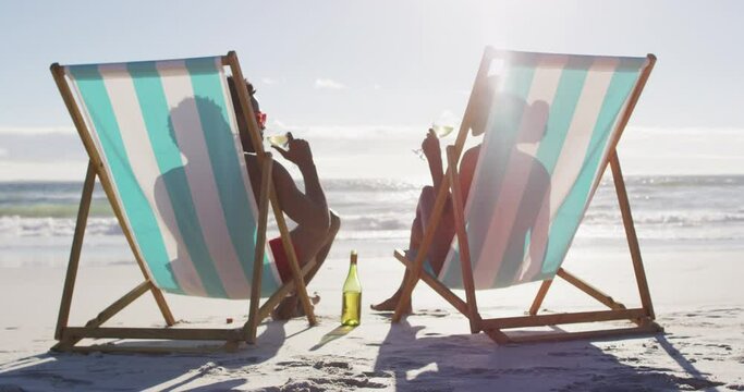 African american couple drinking wine together sitting on deck chairs at the beach
