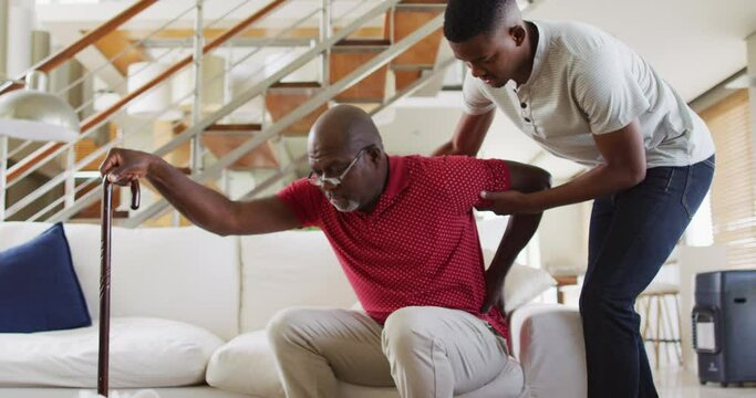 African american senior father getting up the couch and using a cane with teenage son helping