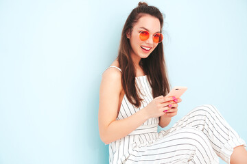Naklejka premium Young beautiful smiling female in trendy summer hipster overalls clothes. Sexy carefree woman posing near light blue wall in studio. Positive model having fun indoors. Enjoying using smartphone apps