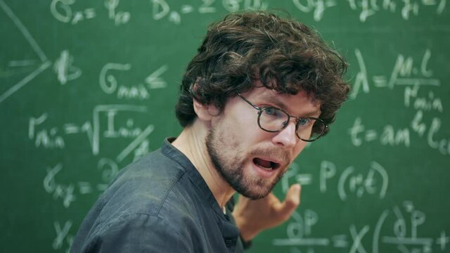 Portrait middle aged stylish teacher talks explains mathematical formulas that written on blackboard behind him. Male lecturer communicates with students in university classroom