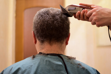 A man is trimmed with a hair clipper and sits with his back to the camera. Perhaps at home in a lockdown