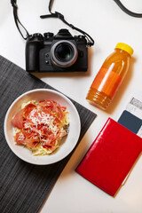 Food at the airport. On the table is a plate of pasta and Parma ham, a camera and juice in bottles. Passport with plane ticket. Travels. What is served at the airports. The mood for new adventures.